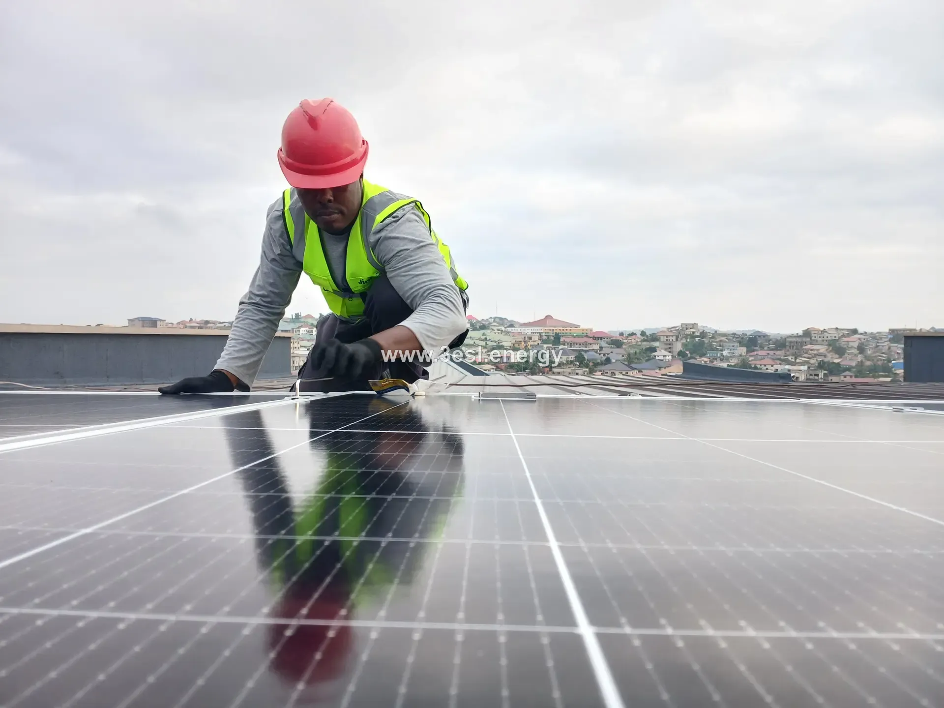 A technician in a red hard hat wiping down a solar module, the sky reflected in its surface.