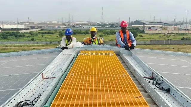 Three workers in hard hats and high-visibility vests kneeling on a yellow rooftop walkway between module rows.
